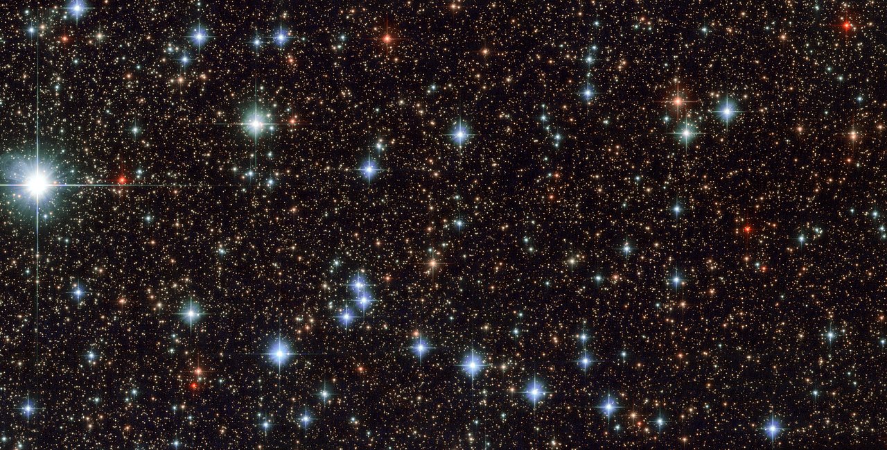 Dicht gedrängtes Sternfeld im Sternbild Schütze, aufgenommen vom Hubble-Weltraumteleskop: Tausende gelbliche, bläuliche und rote Sterne stehen vor dem Band der Milchstraße, links außen leuchtet ein besonders heller Vordergrundstern mit Beugungskreuz.