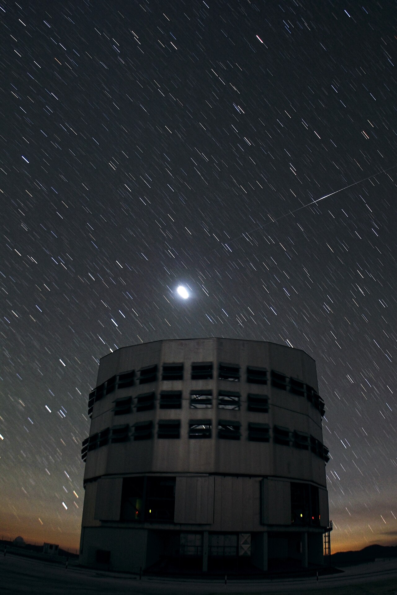 Heller, kurzer Lichtstreifen eines Iridium-Satelliten am Nachthimmel über der Kuppel des Very Large Telescope Yepun