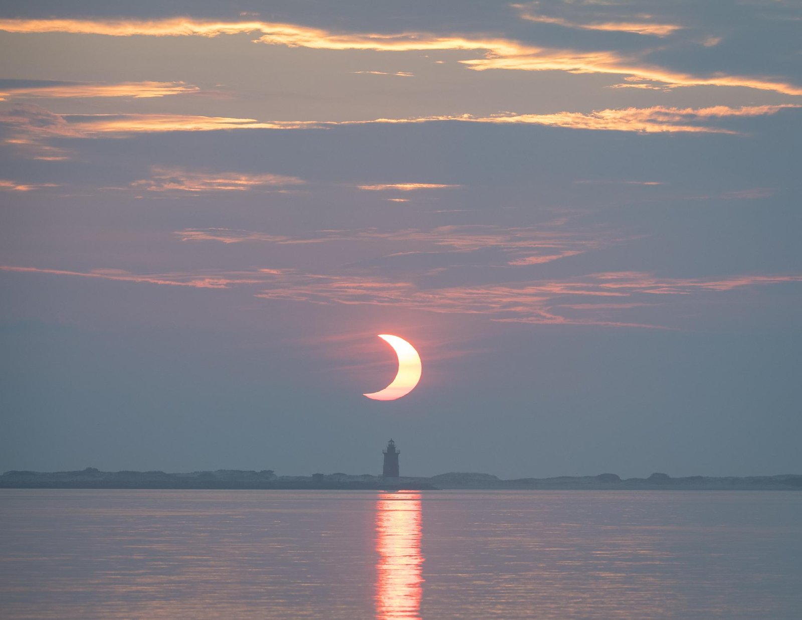 Sichelförmige, teilweise verfinsterte Sonne geht über dem Meer hinter einem Leuchtturm auf